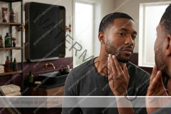 Black man with rich brown skin calmly checking sensitive skin and dark spots in a mirror